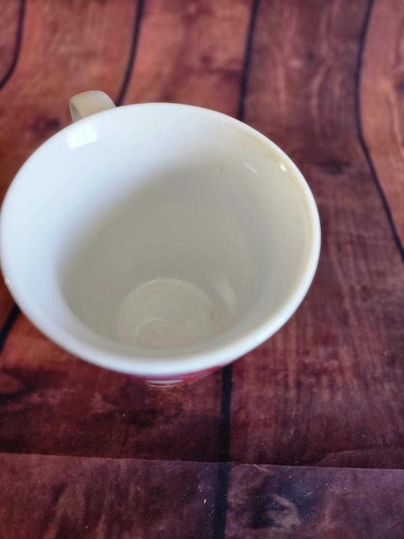 Puede incluir: Una taza de cer&aacute;mica blanca con un asa peque&ntilde;a. El interior de la taza es blanco y el borde tiene un sutil acento rosa. La taza est&aacute; sobre una superficie de madera marr&oacute;n.