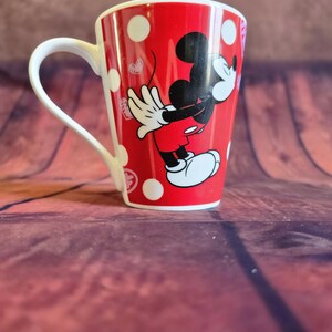 Puede incluir: Taza de cer&aacute;mica roja con lunares blancos y una imagen de dibujos animados de Mickey Mouse. La taza tiene un asa blanca y un interior rojo. Mickey Mouse lleva pantalones cortos rojos, guantes blancos y zapatos blancos.