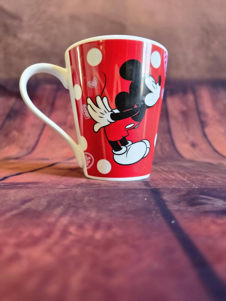 Puede incluir: Taza de cer&aacute;mica roja con lunares blancos y una imagen de dibujos animados de Mickey Mouse. La taza tiene un asa blanca y un interior rojo. Mickey Mouse lleva pantalones cortos rojos, guantes blancos y zapatos blancos.