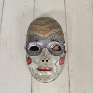 Peut inclure: Un masque artisanal au design unique. Le masque présente une base grise avec des lèvres et des joues rouges. Le front présente un motif en chevrons dans les tons marron et vert, avec des couvre-yeux argentés.
