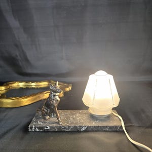 Peut inclure: Une lampe décorative avec un petit abat-jour en verre dépoli et une figurine de chien en métal foncé. La lampe repose sur une base rectangulaire en marbre. Une pièce décorative dorée est en arrière-plan.