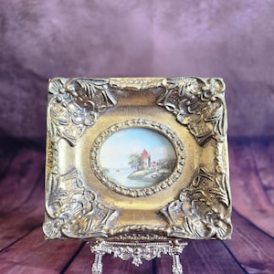 Peut inclure: Un petit cadre doré antique avec une peinture ovale d'un paysage. Le cadre présente des détails floraux ornés et repose sur un chevalet décoratif argenté. La peinture représente un bâtiment près d'un plan d'eau.