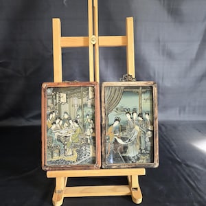 Peut inclure: Deux œuvres d'art rectangulaires encadrées représentant des scènes détaillées de personnages en tenue traditionnelle. Le cadre de gauche montre un groupe autour d'une table, tandis que celui de droite montre des personnages dans un intérieur. Cadres en bois usé.