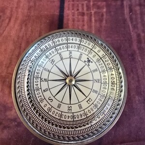 Puede incluir: Una brújula circular de latón con una esfera blanca que muestra los puntos cardinales negros N, NE, E, SE, S, SW, W y NW. El borde exterior presenta una escala numérica detallada del 0 al 90 en ambas direcciones, con un patrón decorativo.