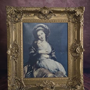 Peut inclure: Impression encadrée dorée ornée d'une mère et son enfant. L'œuvre représente une femme en robe sombre et un enfant en robe claire. Le cadre présente des volutes détaillées et est exposé sur un chevalet en bois.
