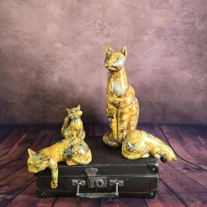 Peut inclure: Quatre figurines décoratives de chats aux tons de jaune, marron et gris sont posées sur une valise vintage marron. Les chats sont dans différentes poses : debout, assis et couchés. L'arrière-plan est marron texturé.