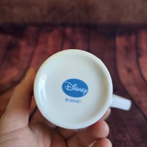 Puede incluir: Taza de cer&aacute;mica blanca con asa, con un logotipo ovalado azul con la palabra "Disney" y el s&iacute;mbolo de copyright. La taza se sostiene en una mano, con un fondo de madera.