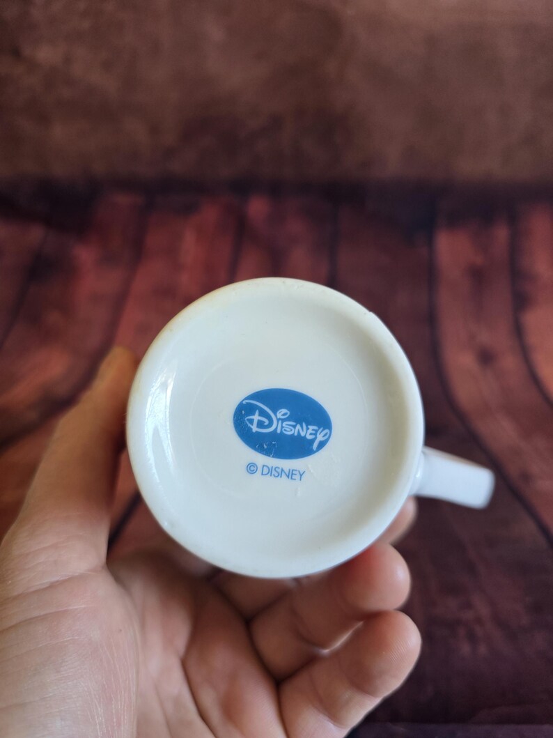 Puede incluir: Taza de cer&aacute;mica blanca con asa, con un logotipo ovalado azul con la palabra "Disney" y el s&iacute;mbolo de copyright. La taza se sostiene en una mano, con un fondo de madera.