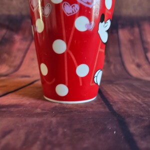 Puede incluir: Un vaso rojo con lunares blancos y dise&ntilde;os en forma de coraz&oacute;n. El vaso presenta la cara de un personaje de dibujos animados. Es cil&iacute;ndrico con un borde blanco, sobre un fondo de madera marr&oacute;n.