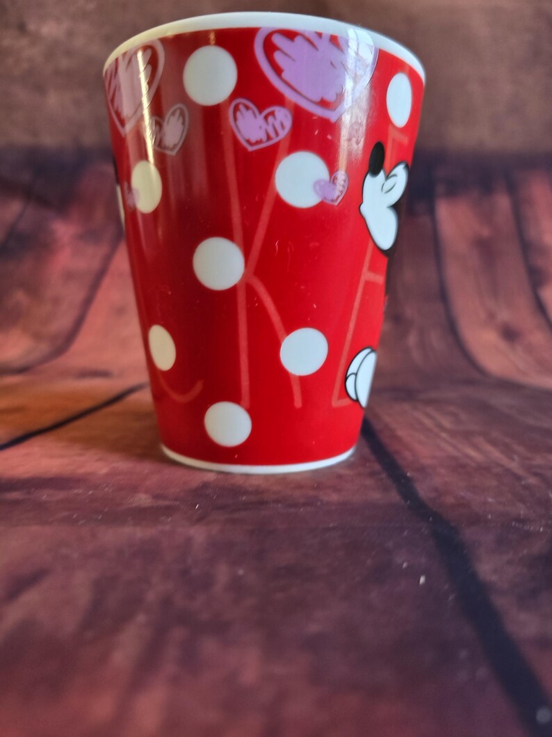 Puede incluir: Un vaso rojo con lunares blancos y dise&ntilde;os en forma de coraz&oacute;n. El vaso presenta la cara de un personaje de dibujos animados. Es cil&iacute;ndrico con un borde blanco, sobre un fondo de madera marr&oacute;n.