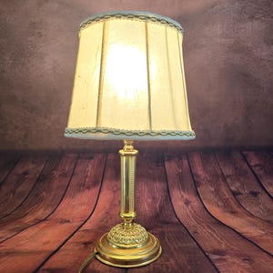 May include: A vintage table lamp with a gold-colored base and stem, topped with a cream-colored lampshade. The lampshade is trimmed with a delicate, light blue lace. The lamp is plugged in and lit, casting a warm glow.