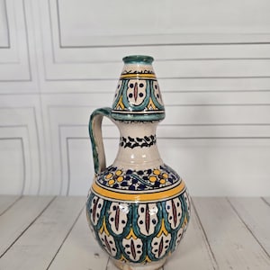Peut inclure: Une cruche en céramique avec une anse, ornée de motifs floraux turquoise, jaunes et bordeaux sur une base blanche. La cruche a un col étroit et un petit bec, avec un motif assorti sur le dessus. La poignée est turquoise.