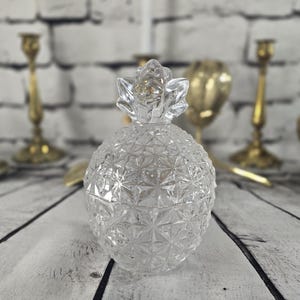 Peut inclure: Un pot en verre transparent en forme d'ananas avec un couvercle. Le pot est posé sur une surface en bois blanc, avec des chandeliers dorés flous et d'autres objets décoratifs en arrière-plan. Le pot est un objet décoratif.