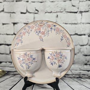 Peut inclure: Poche murale en céramique crème avec deux jardinières en forme de coupe. La pièce est décorée de motifs floraux roses et bleus. La plaque arrière ronde a une bordure florale. La poche murale est exposée sur un support noir.