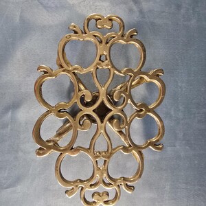 May include: An ornate gold-coloured metal trivet with an openwork design. The trivet features a symmetrical pattern of interconnected shapes, including stylized apple-like forms. The metal has a polished finish. Designed to protect surfaces from hot cookware.