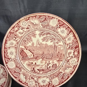 Peut inclure: Ensemble d'assiettes anciennes en céramique rouge et blanche. Chaque assiette présente une scène de paysage centrale avec un pont, des bâtiments et des personnages dans un bateau. La bordure est ornée de motifs floraux. Les assiettes sont présentées sur un support doré.