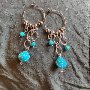 May include: Silver hoop earrings with turquoise and white accents. The earrings feature small turquoise beads, silver chain links, and larger turquoise and white patterned beads at the bottom. The earrings are displayed against a textured, gray-green background.
