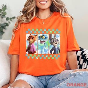 May include: Orange t-shirt featuring three cartoon characters in a checkered frame. The characters include a fox in a pink shirt, a blue snake, and a rabbit in a blue jacket. The word "ORANGE" is printed at the bottom of the shirt.