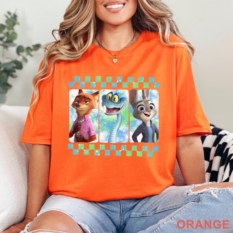 May include: Orange t-shirt featuring three cartoon characters in a checkered frame. The characters include a fox in a pink shirt, a blue snake, and a rabbit in a blue jacket. The word "ORANGE" is printed at the bottom of the shirt.