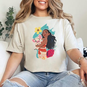 May include: Beige t-shirt featuring a graphic of a Polynesian woman holding a pig. The woman has long black hair and is wearing a red and white outfit. A yellow flower and blue accents are in the background.