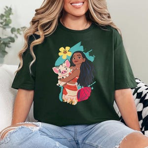 May include: Dark green t-shirt featuring a graphic of a woman holding a pig, with a flower and seashell design. The woman has long black hair and is wearing a red and white outfit. The shirt is a casual style.