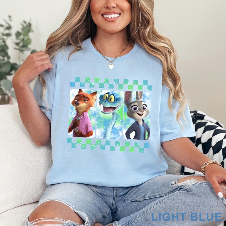 May include: Light blue t-shirt featuring three cartoon characters in a checkered frame. The characters include a fox in a pink shirt, a blue snake, and a rabbit in a blue jacket. The text "LIGHT BLUE" is at the bottom.