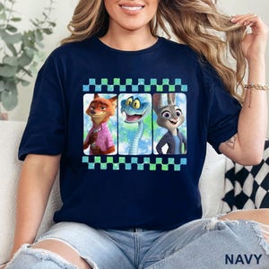 May include: Navy blue t-shirt featuring three characters from Zootopia: Nick Wilde, a snake, and Judy Hopps. The characters are in individual frames with a checkered border. The word "NAVY" is printed at the bottom of the shirt.