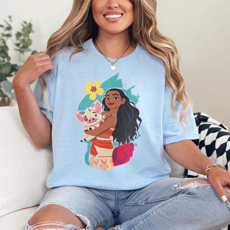 May include: Light blue t-shirt featuring a graphic of a woman holding a pig, with a yellow flower and teal brushstrokes. The woman has long black hair and is wearing a red top and skirt. A red seashell is also visible.
