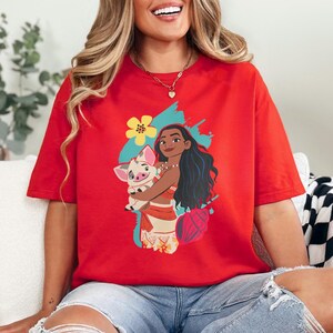 May include: Red t-shirt featuring a cartoon illustration of a woman with long black hair holding a pig. The graphic includes a yellow flower, a blue splash, and a pink seashell. The woman is wearing a red and tan outfit.