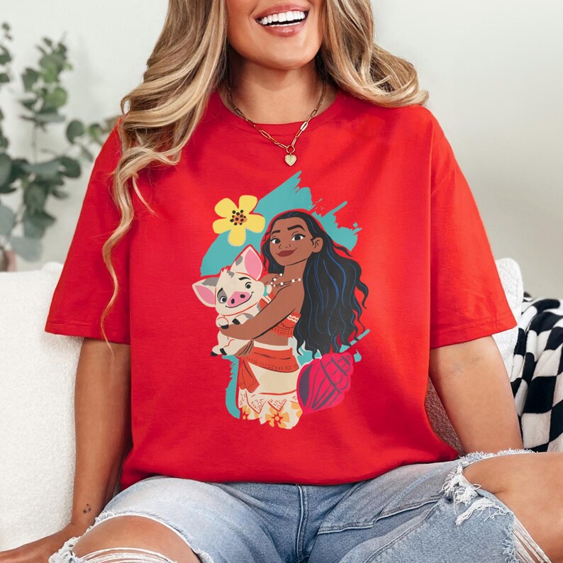 May include: Red t-shirt featuring a cartoon illustration of a woman with long black hair holding a pig. The graphic includes a yellow flower, a blue splash, and a pink seashell. The woman is wearing a red and tan outfit.
