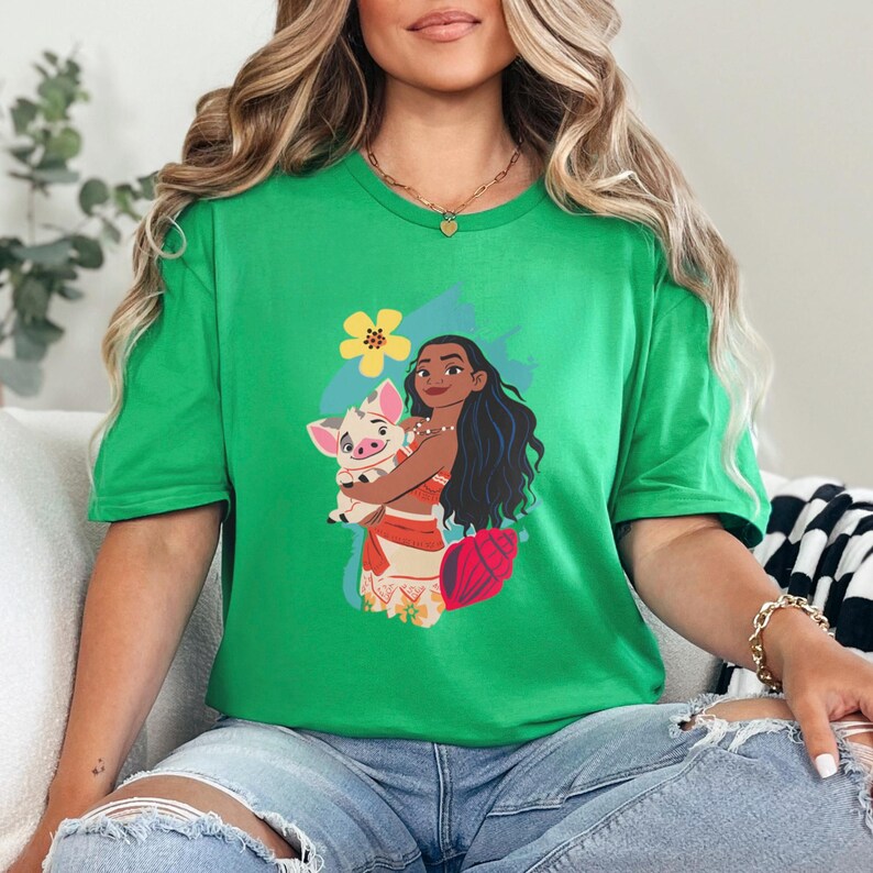 May include: A bright green t-shirt featuring a graphic of a cartoon character holding a pig. The character has long black hair and is wearing a red and orange outfit. A yellow flower and blue background are also present.