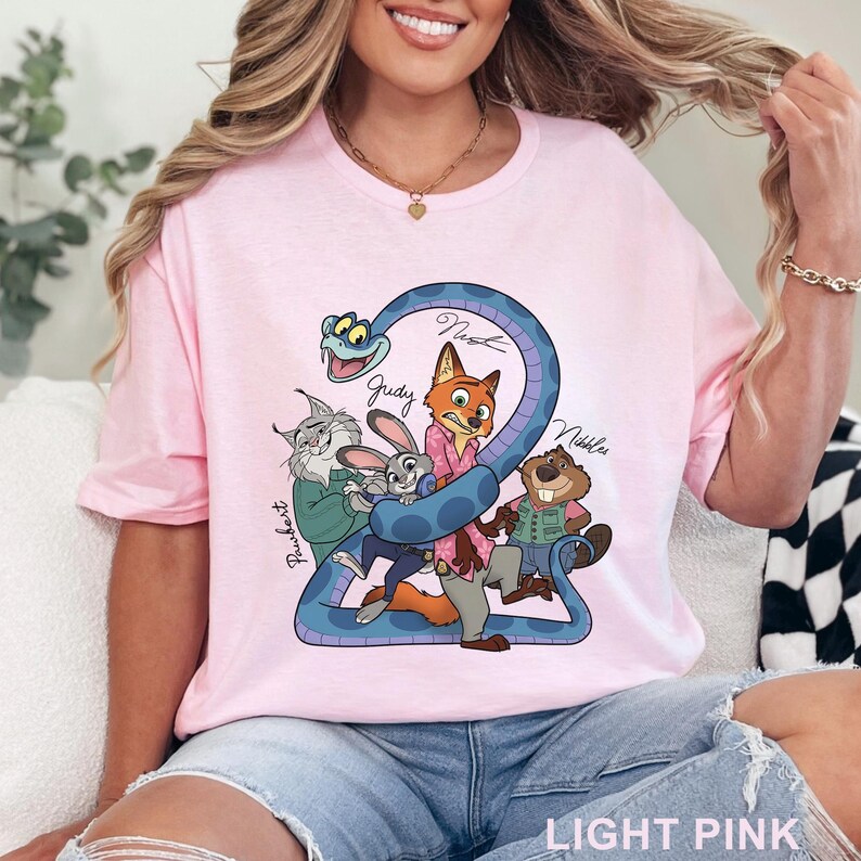 May include: Light pink t-shirt featuring a cartoon graphic of several animal characters, including a fox, rabbit, and beaver, with the names "Judy", "Nick", and "Nibbles" written on the design. The text "LIGHT PINK" is printed at the bottom.