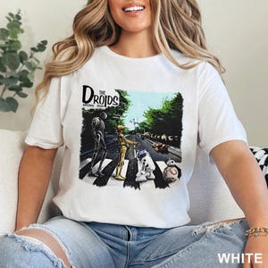 May include: White t-shirt featuring a graphic of Star Wars droids crossing a street, a parody of the Beatles' Abbey Road album cover. The text "The Droids Imperial Road" is above the image. The shirt is labeled "WHITE" at the bottom.