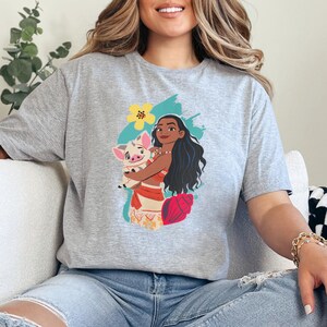May include: A gray t-shirt featuring a colorful graphic of a woman with long black hair holding a pig. The graphic includes a yellow flower, a seashell, and a turquoise background. The woman is wearing a red and orange outfit.