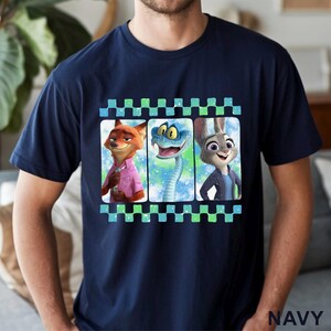 May include: Navy blue t-shirt featuring a graphic of three animated characters: a fox, a snake, and a rabbit. The characters are set against a colorful background within a film strip design. The word "NAVY" is printed at the bottom.