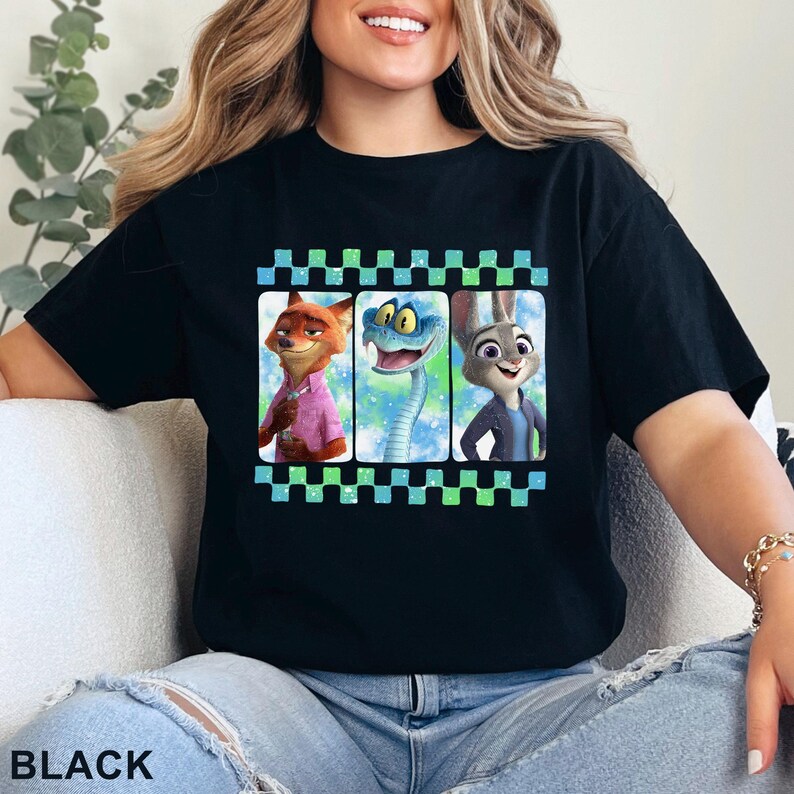 May include: Black t-shirt featuring a colorful graphic of three animated characters: a fox in a pink shirt, a snake, and a rabbit. The design is framed by a checkered pattern in green and blue. The word "BLACK" is printed at the bottom.