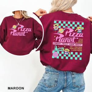A person wearing a maroon sweatshirt with a colorful, retro-style design featuring characters from the movie "Pizza Planet".