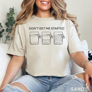 May include: Sand-colored t-shirt with the text "DON'T GET ME STARTED" above three jars. The jars have varying levels of contents, with bubbles in the last two. Each jar has a small tag. The word "SAND" is at the bottom.