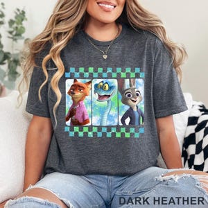 May include: Dark heather gray t-shirt featuring a graphic of three cartoon characters: a fox, a snake, and a rabbit. The graphic is framed with a checkered pattern in blue and green. The text "DARK HEATHER" is printed at the bottom.
