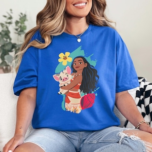 May include: Royal blue t-shirt featuring a graphic of a cartoon woman holding a pig. The woman has long black hair and is wearing a red and white outfit. A yellow flower and blue paint strokes are behind the characters.