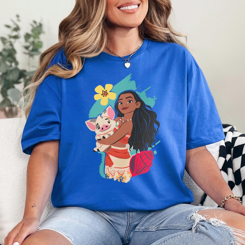 May include: Royal blue t-shirt featuring a graphic of a cartoon woman holding a pig. The woman has long black hair and is wearing a red and white outfit. A yellow flower and blue paint strokes are behind the characters.