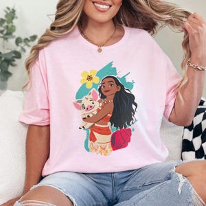 May include: A light pink t-shirt featuring a graphic of a woman with long black hair holding a pig. The graphic includes a yellow flower, a teal brushstroke, and a red seashell. The woman is wearing a red top and a skirt with a floral pattern.