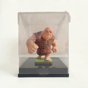 Op de afbeelding: Een kaal, bebaard figuurtje met oranje gezichtshaar dat een bruine tuniek met riem draagt, staand op een groene basis in een doorzichtige plastic displaydoos met een zwarte vierkante bodem. Het personage heeft een gespierde bouw en is blootsvoets.