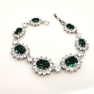 May include: A silver bracelet with a row of emerald green oval-shaped stones surrounded by smaller clear stones.