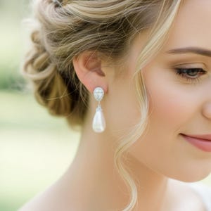 Puede incluir: Primer plano de una mujer con elegantes pendientes colgantes. Los pendientes tienen un colgante de perla en forma de lágrima y un detalle redondo y brillante. Su cabello rubio está peinado en un moño con rizos sueltos. Los pendientes están engarzados en metal plateado.