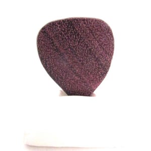 May include: A dark purple, heart-shaped guitar pick with a textured surface. The pick is set against a white background, highlighting its shape and color. This accessory is designed for guitar players.