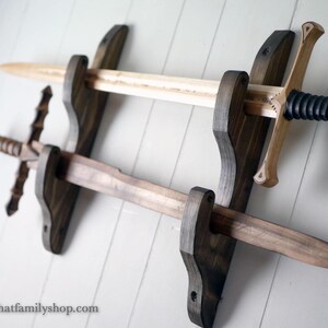 Puede incluir: Un soporte de pared de madera teñida oscura que sostiene dos espadas de madera. La espada superior es de color claro con una empuñadura envuelta en negro, mientras que la inferior es de madera marrón oscura con una guarda texturizada. El texto thatfamilyshop.com es visible.