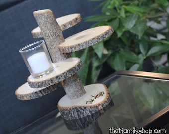 May include: A wooden candle holder with a tree trunk design and four tiers. The holder is decorated with bark and has the words "Tree Logs" engraved on the bottom tier. A small glass candle holder sits on the top tier.