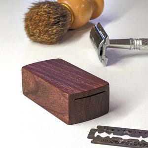 Puede incluir: Una caja rectangular de madera oscura para guardar cuchillas de afeitar, una maquinilla de afeitar de metal plateado con mango texturizado, una brocha de afeitar de madera con cerdas marrones y una cuchilla de acero inoxidable con texto.