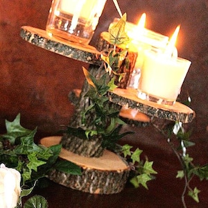 May include: A tiered candle holder made from natural wood slices with bark edges, featuring three clear glass votive cups containing lit candles. The structure is decorated with green artificial ivy vines and a single white rose sits nearby. Text reads Home'nStead.